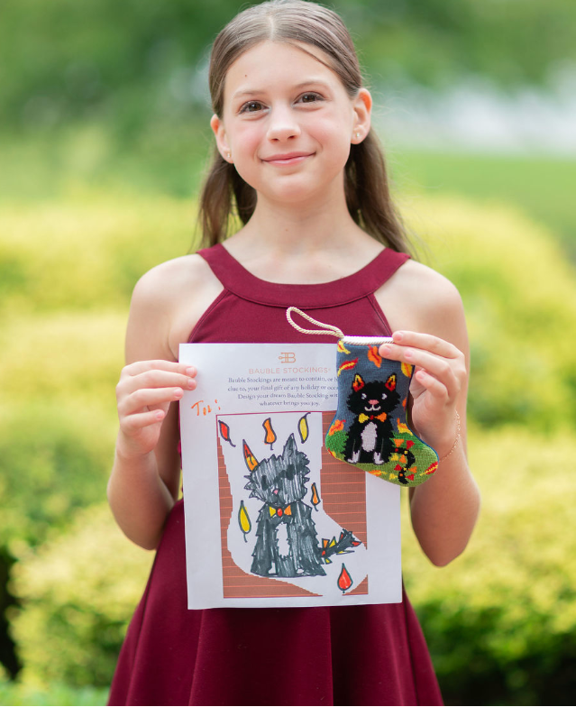 A young girl with long brown hair stands outdoors in a garden setting, holding up a printed sewing pattern booklet and a small handmade black cat Bauble Stocking with colorful embroidered details.