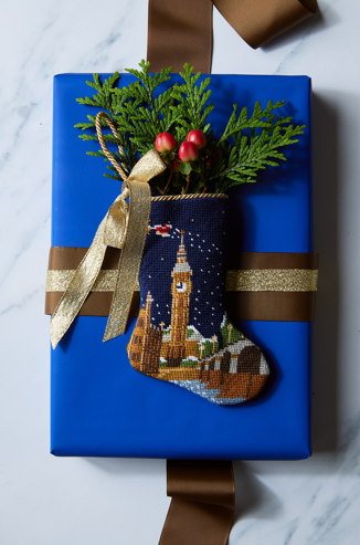 Hand-stitched needlepoint stocking styled on a blue gift box with festive greenery sprig