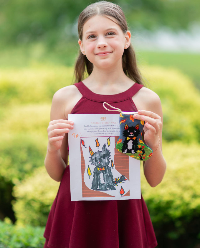 A young girl with brown hair in a ponytail, wearing a sleeveless burgundy dress, holds up a completed craft kit. The kit includes a printed instruction sheet showing a castle design with flames and a small black cat bead ornament.