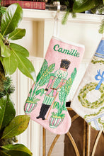 Full-size Christmas stocking with a monogrammed name in script featuring a vibrant nutcracker with festive details and palm accents on pink background