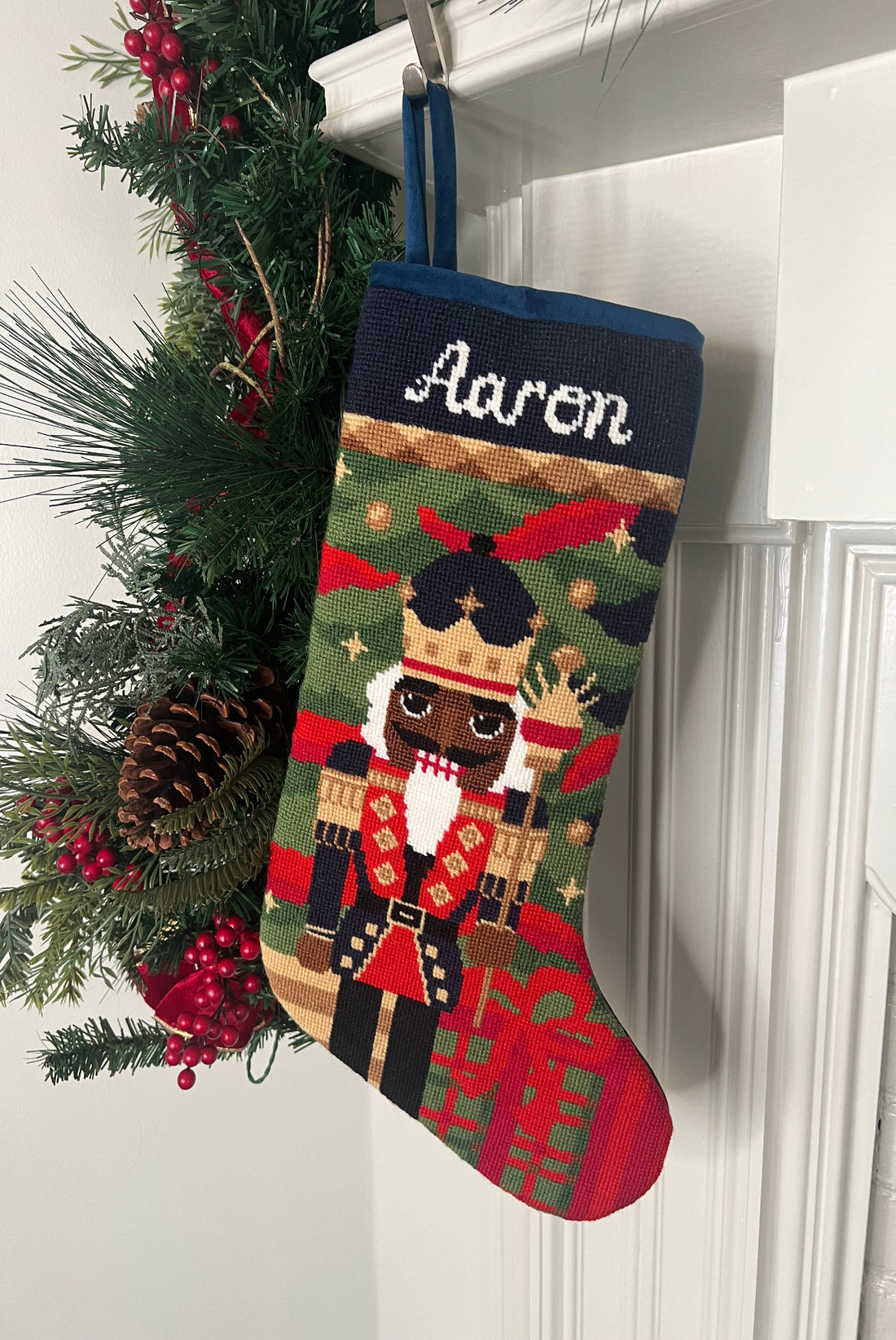 Full-size Christmas stocking with a monogrammed name in script featuring a Nutcracker wearing a vibrant uniform and a golden crown in a greenery background with gift boxes.