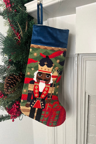 Full-size Christmas stocking featuring a Nutcracker in a vibrant uniform and golden crown, set against a backdrop of greenery and gift boxes.