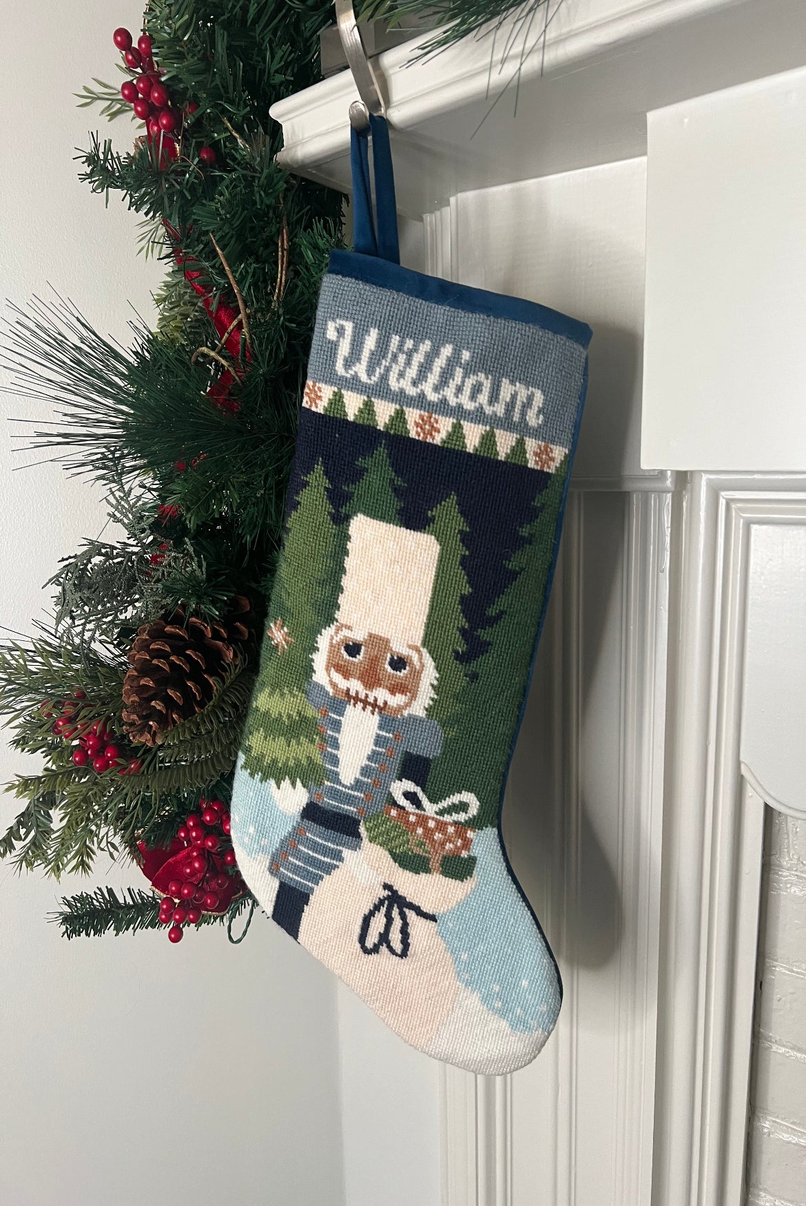 Full-size Christmas stocking with a monogrammed name in script featuring a Nutcracker holding a sack of gifts, standing on snowy ground, with trees in the background
