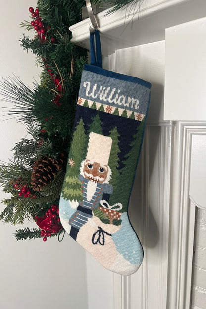Full-size Christmas stocking with a monogrammed name in script featuring a Nutcracker holding a sack of gifts, standing on snowy ground, with trees in the background