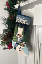 Full-size Christmas stocking with a monogrammed name in script featuring a Nutcracker holding a sack of gifts, standing on snowy ground, with trees in the background