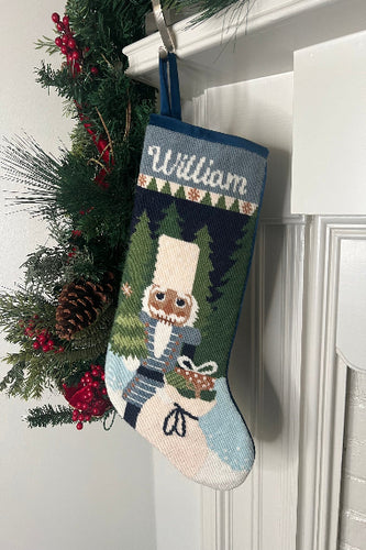 Full-size Christmas stocking with a monogrammed name in script featuring a Nutcracker holding a sack of gifts, standing on snowy ground, with trees in the background