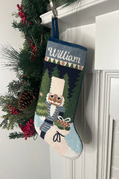 Full-size Christmas stocking with a monogrammed name in script featuring a Nutcracker holding a sack of gifts, standing on snowy ground, with trees in the background