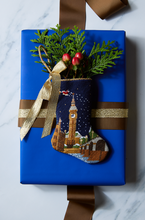 Hand-stitched needlepoint stocking styled on a blue gift box with festive greenery sprig