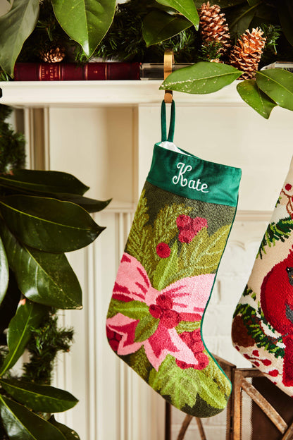 Full-size Christmas stocking with a monogrammed name in script featuring a pink bow with holly and pine branches on a green backdrop