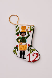 Hand-stitched Christmas stocking featuring drummer in green coat with yellow hat surrounded by greenery on white background