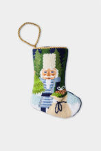 Christmas stocking featuring a Nutcracker holding a sack of gifts, standing on snowy ground, with trees in the background.